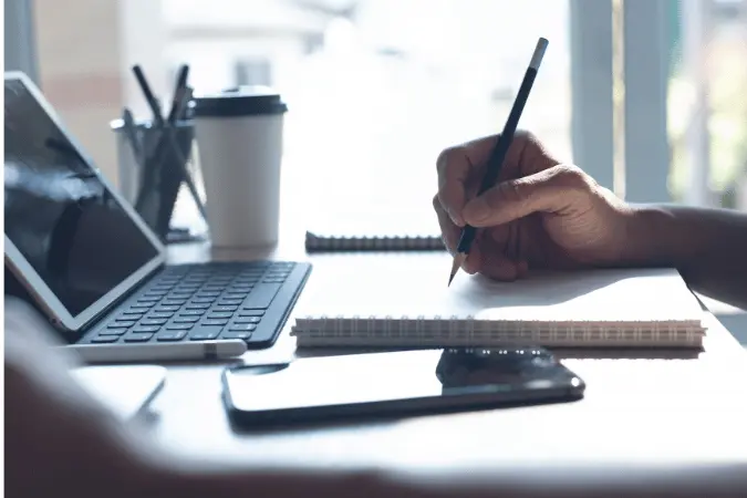 Person writing notes while studying on a laptop at a desk with coffee and phone nearby