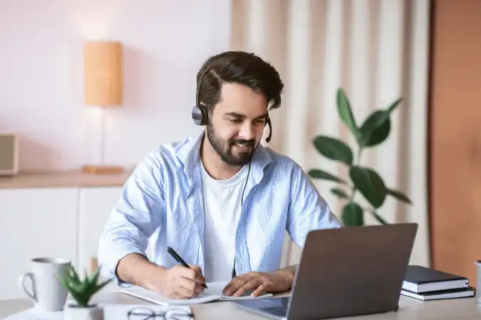 Man wearing a headset taking notes while watching a video on his laptop.