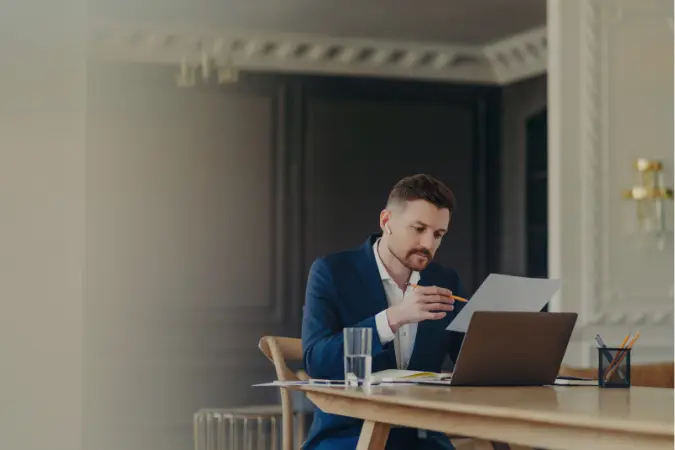 Professional reviewing a document at a desk with a laptop and papers.