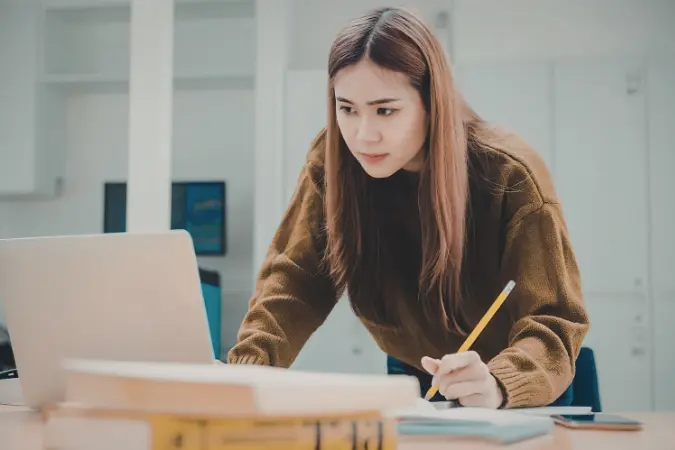Learner reading closely with a pencil in hand beside a laptop and stacked books