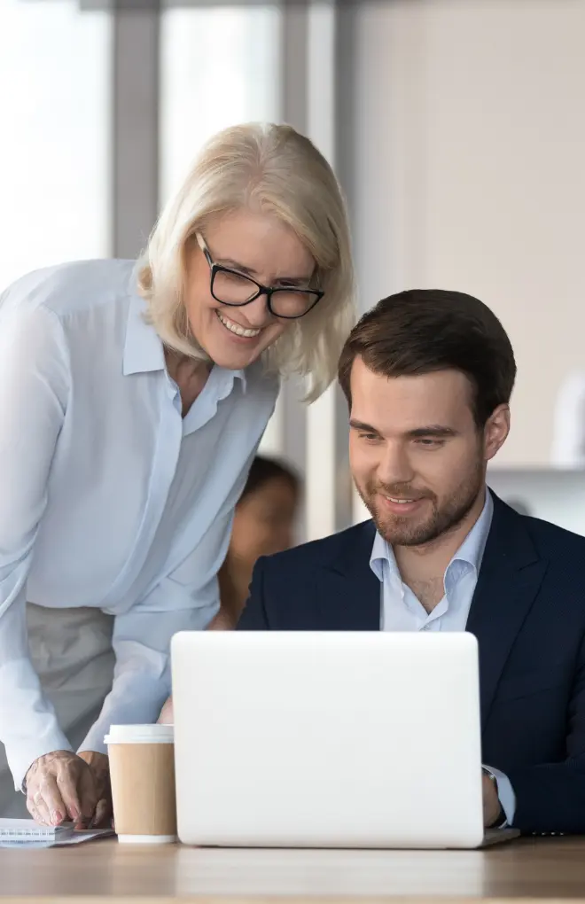 Manager standing behind a seated colleague as they review information on a laptop together in an office.