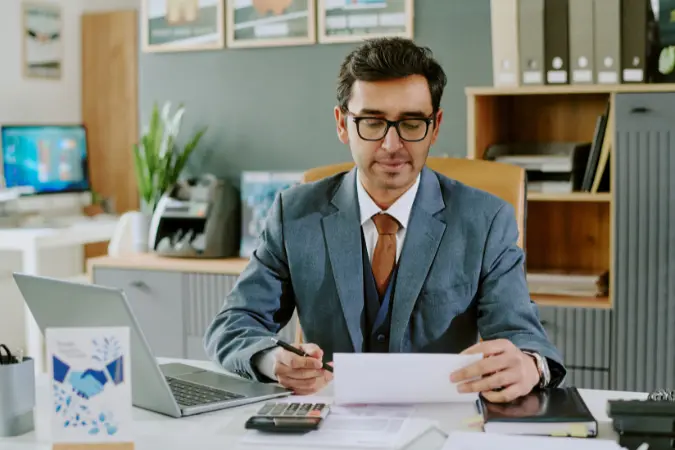 Professional at a desk reading a document with a laptop open beside him.