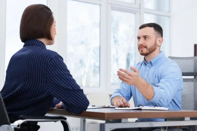 Two professionals in a consultative conversation across a desk in an office.
